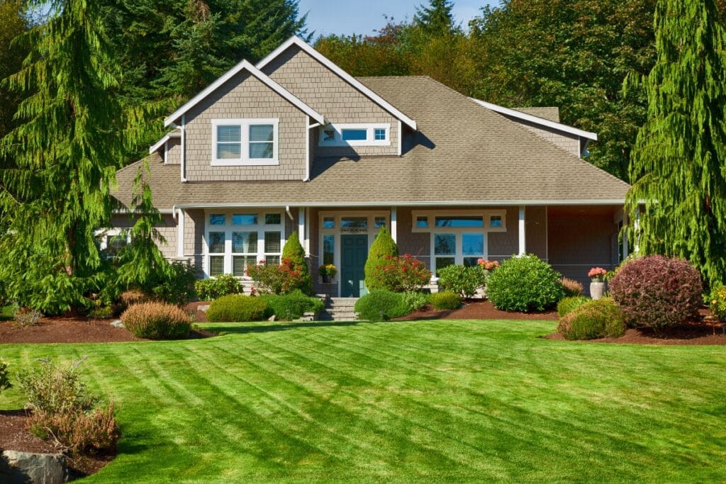 Two-story house with gray siding, white trim, and a covered front porch, surrounded by a landscaped yard featuring Luxury Landscape Design in Georgia with lush green lawn, shrubs, and mature trees.