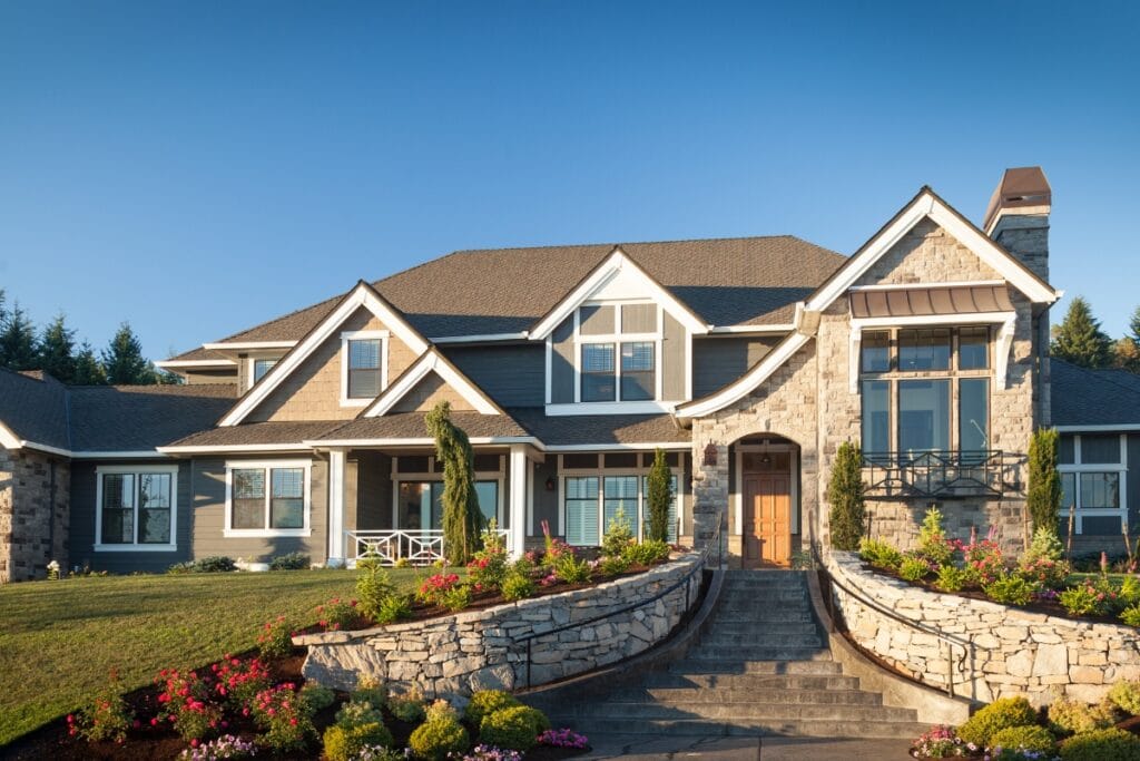 Large two-story house with stone and siding exterior, multiple gables, a central wooden door, manicured lawn, and Luxury Landscape Design in Georgia featuring landscaped flower beds flanking a curved staircase.