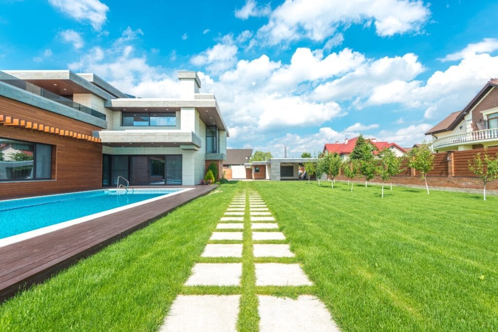 Modern house with large windows, an outdoor swimming pool, manicured green lawn, and stone pathway—showcasing Luxury Landscape Design in Georgia under a blue sky with clouds.