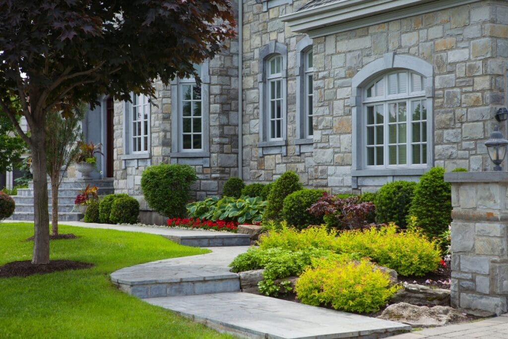 Stone house exterior with arched windows, curved stone walkway, manicured bushes, and colorful flower beds in the front yard—a stunning example of luxury landscape design in Georgia.