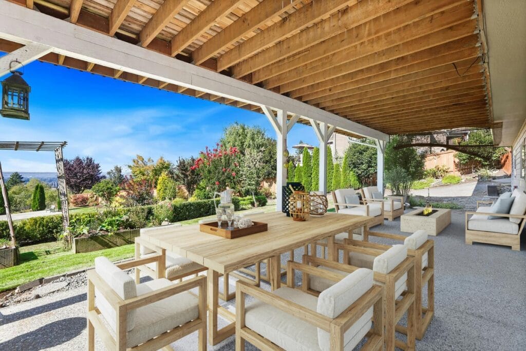 A covered patio with a wooden dining set and lounge seating overlooks a landscaped garden and lawn, showcasing luxury landscape design in Georgia under a clear blue sky.