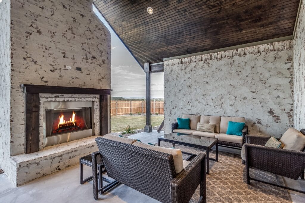 Covered patio with wicker furniture, an inviting outdoor fireplace with a crackling fire, and a view of a grassy backyard enclosed by a wooden fence.