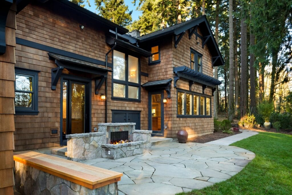 Two-story wooden house with large windows, stone patio, outdoor fireplace, built-in firepit, and surrounding trees, photographed in natural daylight.