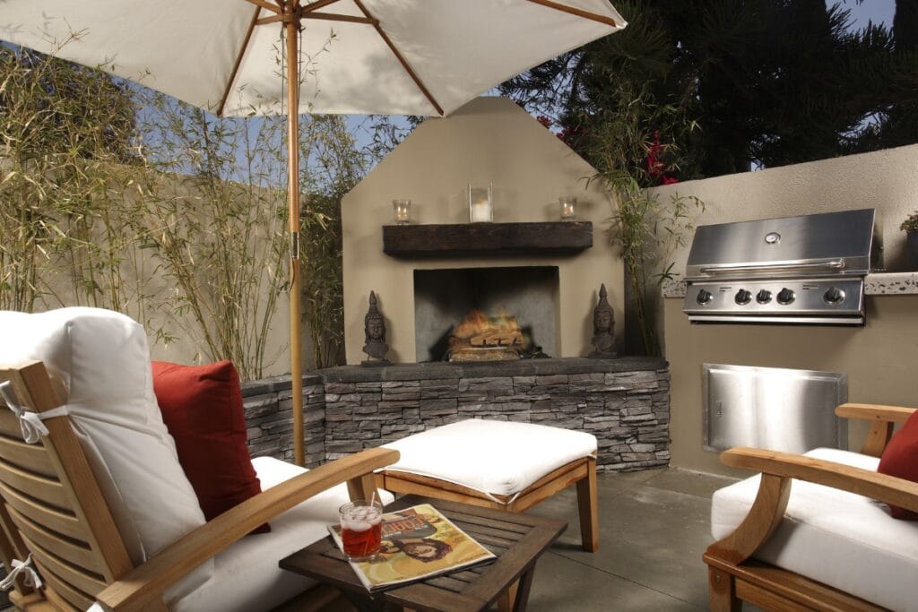 Outdoor patio with cushioned chairs, a small table with a drink and magazine, outdoor fireplaces in stone, built-in grill, and a large umbrella.