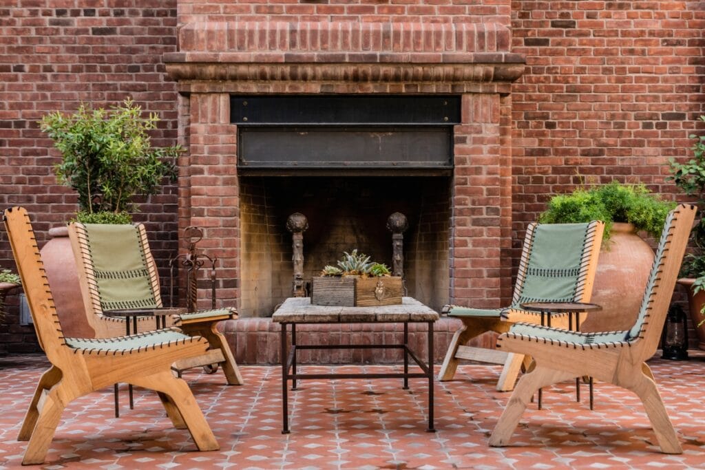 Four wooden chairs with woven backs surround a small table in front of an outdoor fireplace, with potted plants on either side, all set on a tiled floor.