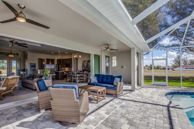 Covered patio with blue cushioned wicker furniture creates an inviting outdoor living space that flows into a modern living room and kitchen; screened enclosure overlooks a backyard and pool.