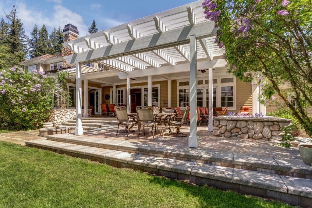 A spacious backyard patio with a pergola, outdoor dining set, stone bar, and seating area creates an inviting Outdoor Living Space, surrounded by greenery and flowering bushes.