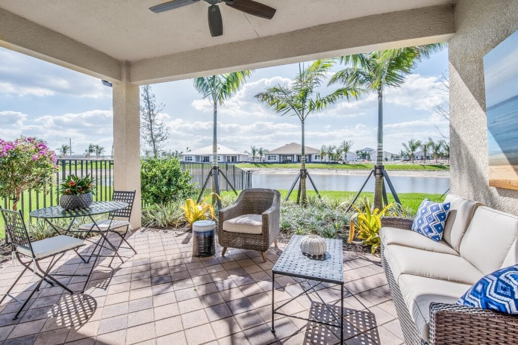 Covered patio with outdoor furniture and potted plants, creating an inviting outdoor living space with a view of a pond and palm trees, all under a ceiling fan on a sunny day.