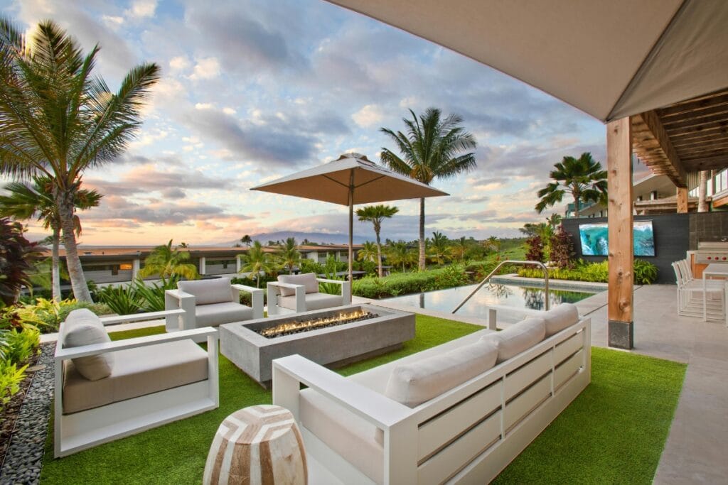 Modern outdoor living space with white furniture, fire pit, umbrella, and a view of the pool and palm trees beneath a partly cloudy sunset sky.