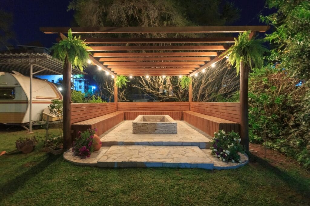 A wooden pergola with string lights, benches, and a central stone firepit creates a cozy outdoor living space on a paved area surrounded by plants at night.