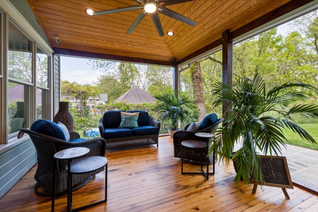 This outdoor living space features a covered patio with wicker seating, blue cushions, a ceiling fan, potted plants, and a wooden floor overlooking a lush green backyard.