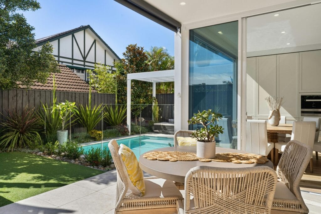 Modern patio with wicker dining set overlooks a backyard pool, creating an inviting outdoor living space surrounded by greenery and a neighboring Tudor-style house. Glass doors seamlessly connect the patio to the indoor kitchen area.