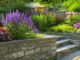 Stone steps and retaining walls border garden beds filled with blooming purple, yellow, and pink flowers, all basking in bright sunlight.