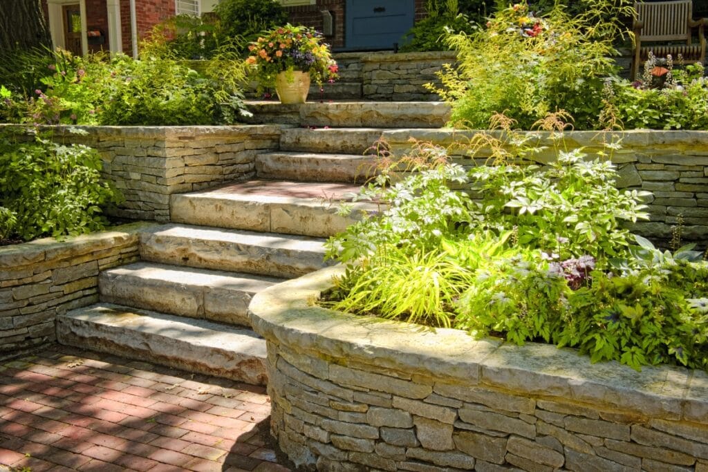 Stone steps and elegant retaining walls border lush green plants and flowers in a beautifully landscaped garden, guiding you toward the house entrance.