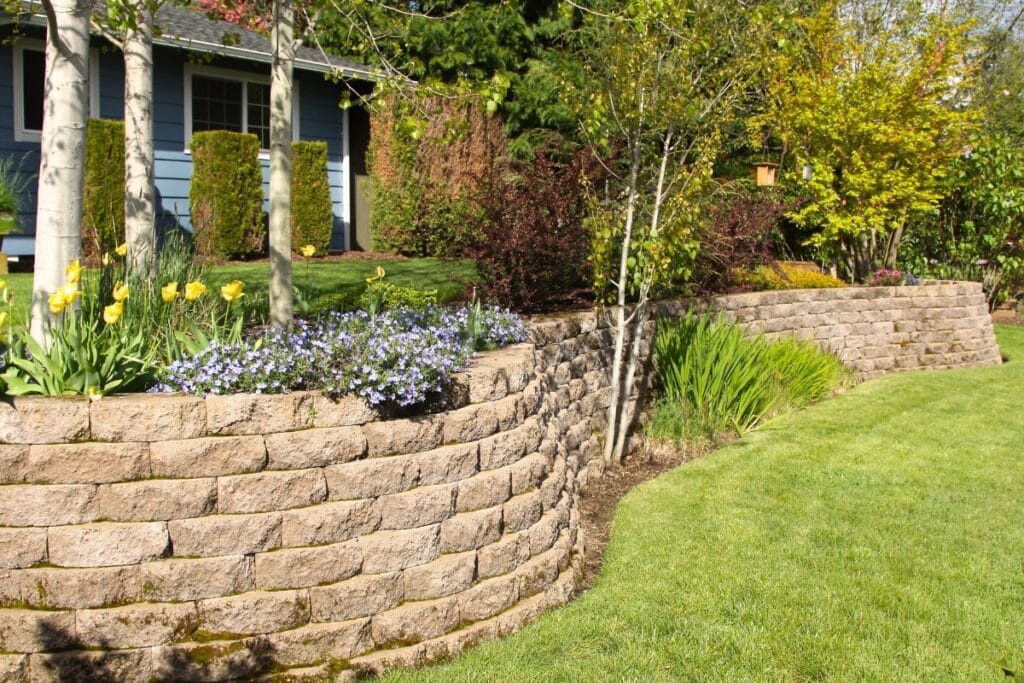A landscaped yard features a curved stone retaining wall, flower beds with yellow tulips and purple blooms, shrubs, and a well-manicured lawn—showcasing the elegance and function of expertly built retaining walls.