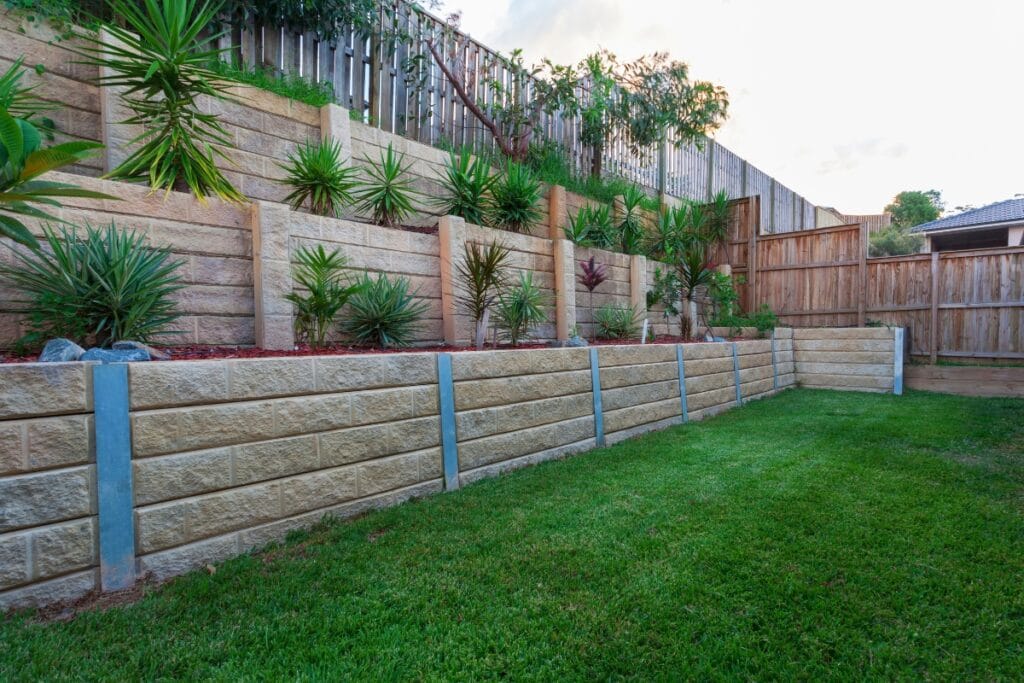 Tiered retaining walls with stone blocks and metal supports hold a landscaped garden filled with various green plants, while wooden fencing surrounds the area.