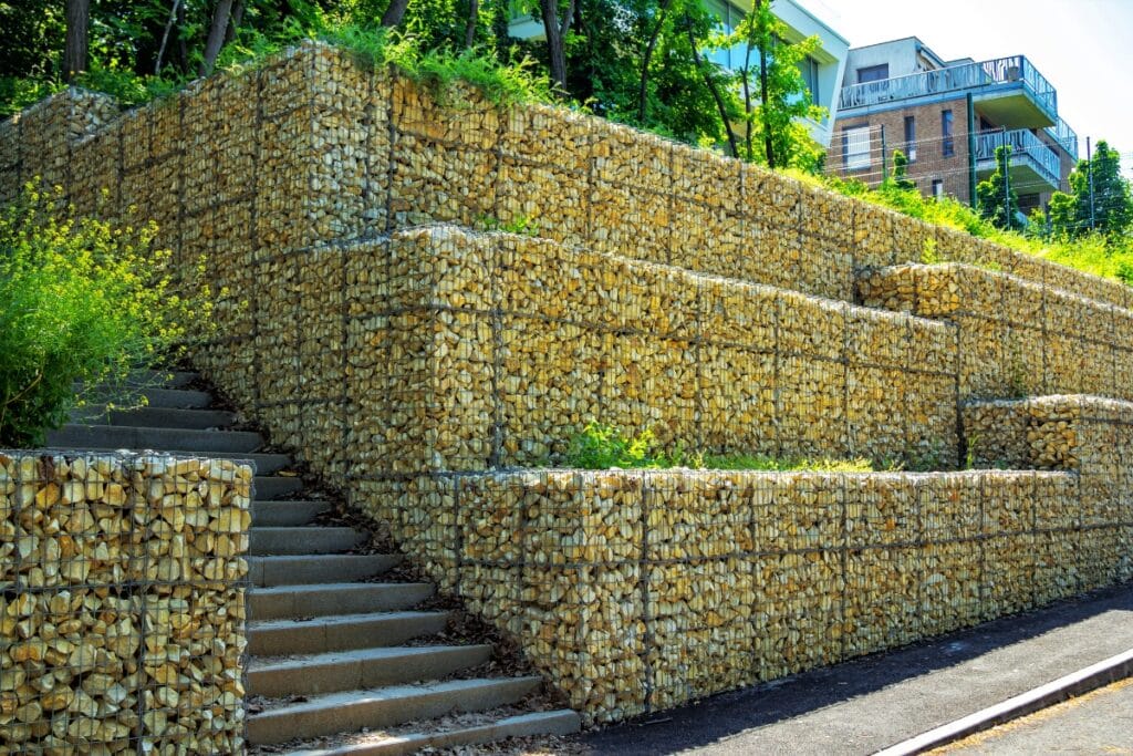 Multi-level retaining walls made from wire mesh filled with rocks, with a concrete staircase built into the structure, surrounded by greenery and modern buildings in the background.