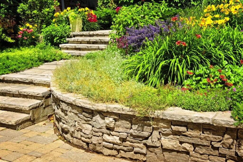 Stone garden steps and a curved retaining wall border a landscaped area filled with colorful flowering plants and lush green foliage, showcasing the beauty and functionality of retaining walls.
