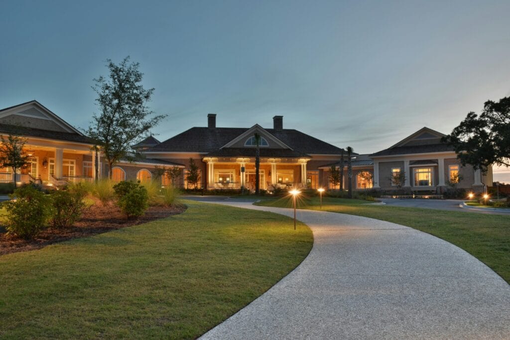 A wide, gently curving pathway with the best outdoor lighting leads to a large, well-lit building with peaked roofs and a landscaped lawn at dusk.