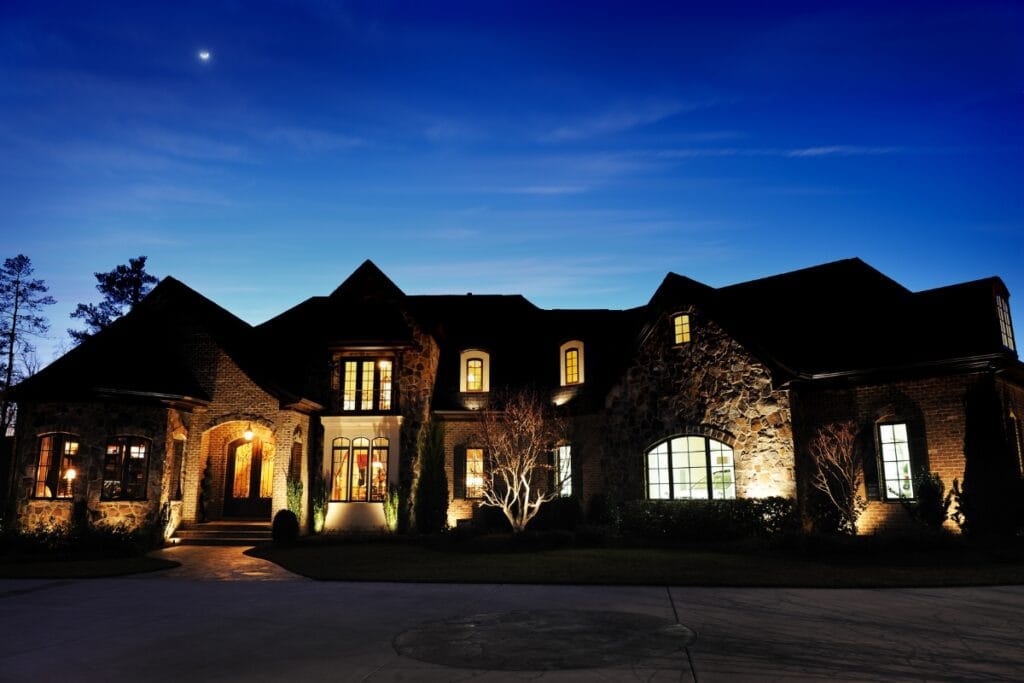 Large stone and brick house illuminated at dusk with the best outdoor lighting, lights glowing from the windows, and a crescent moon visible in the deep blue evening sky.