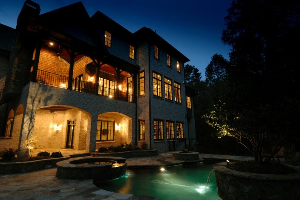 Large brick house at dusk with Best Outdoor Lighting illuminating the windows, a covered balcony, and a backyard featuring a lit swimming pool, hot tub, and stone patio.