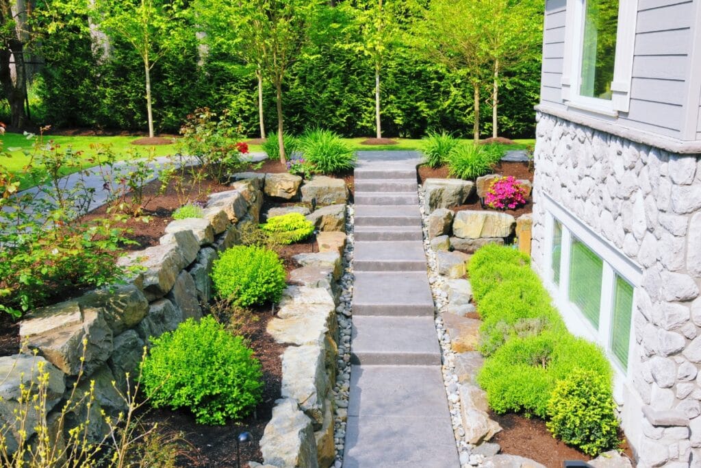 Stone pathway with steps bordered by drought-resistant landscaping—shrubs and rocks—leads up through a landscaped garden to the side of a stone-faced house with large windows.