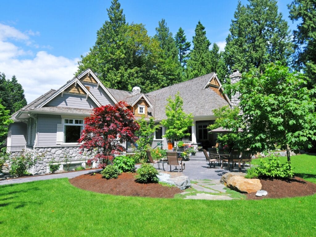 A single-story house with stone and wood siding, surrounded by a well-maintained lawn, trees, shrubs, and a patio with outdoor furniture showcases thoughtful landscape design.