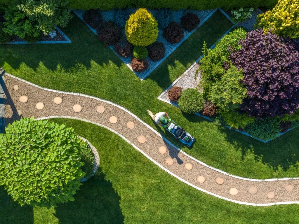 A person mows a neatly manicured lawn along a curved path in a landscaped garden featuring trimmed bushes, decorative gravel, and some of the best trees for shade.