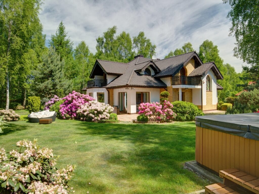 A modern two-story house with a dark roof sits in a lush garden filled with vibrant flowers, shrubs, and the best trees for shade, offering outdoor seating under a partly cloudy sky.