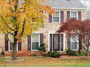 A two-story brick house with dark shutters, a glass front door, and autumn trees and fallen leaves in the front yard, showcasing a healthy lawn with vibrant fall landscaping tips inspired by Cobb County.