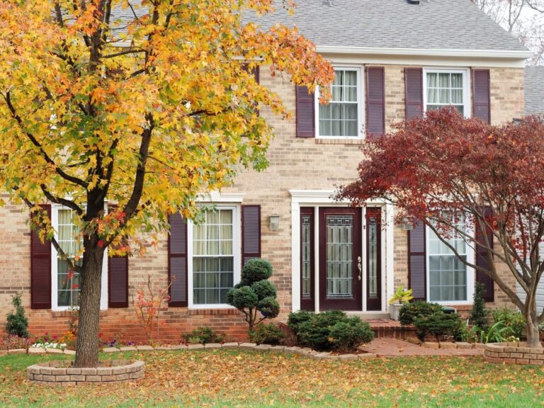 A two-story brick house with dark shutters, a glass front door, and autumn trees and fallen leaves in the front yard, showcasing a healthy lawn with vibrant fall landscaping tips inspired by Cobb County.