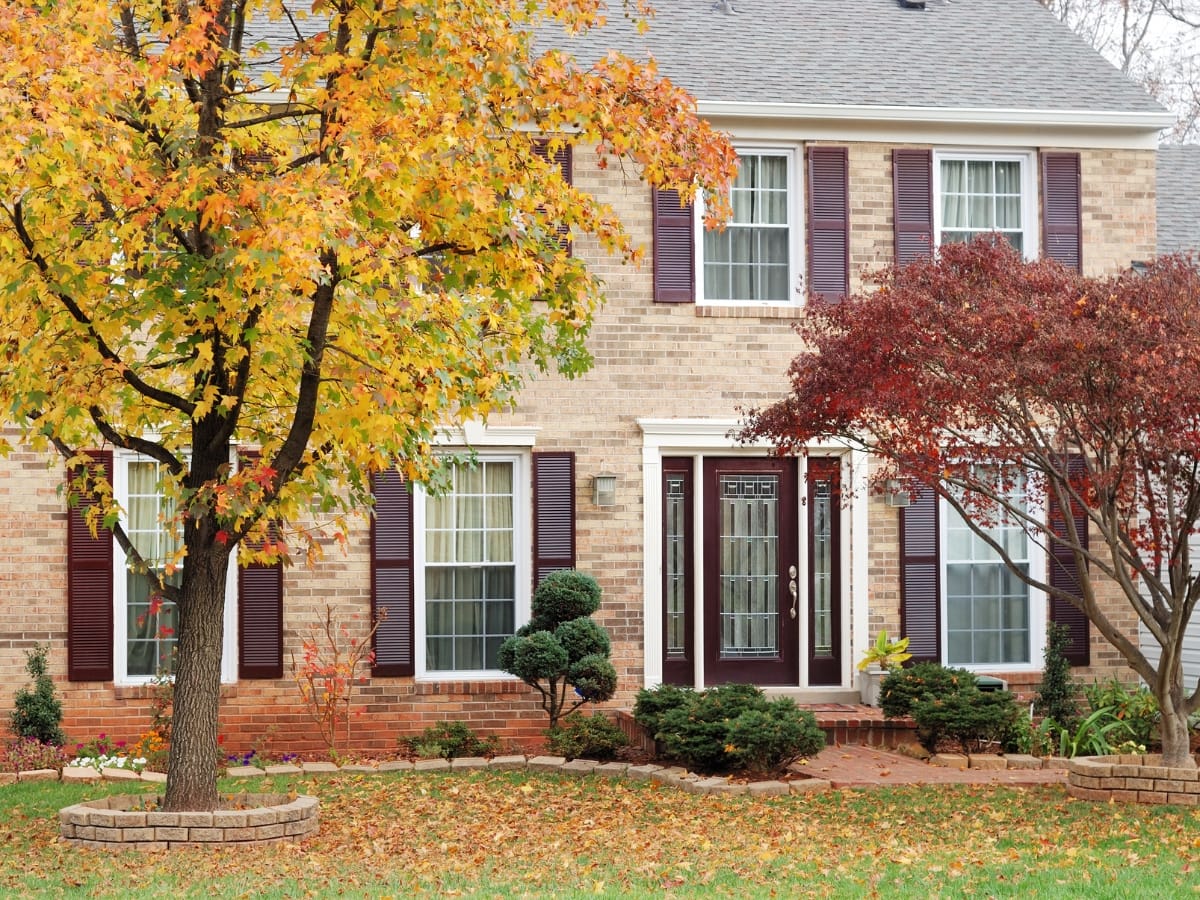 Two-story brick house with maroon shutters and front door, beautifully landscaped yard featuring trees in peak autumn foliage—perfect inspiration for your own home’s curb appeal and fall landscaping tips.