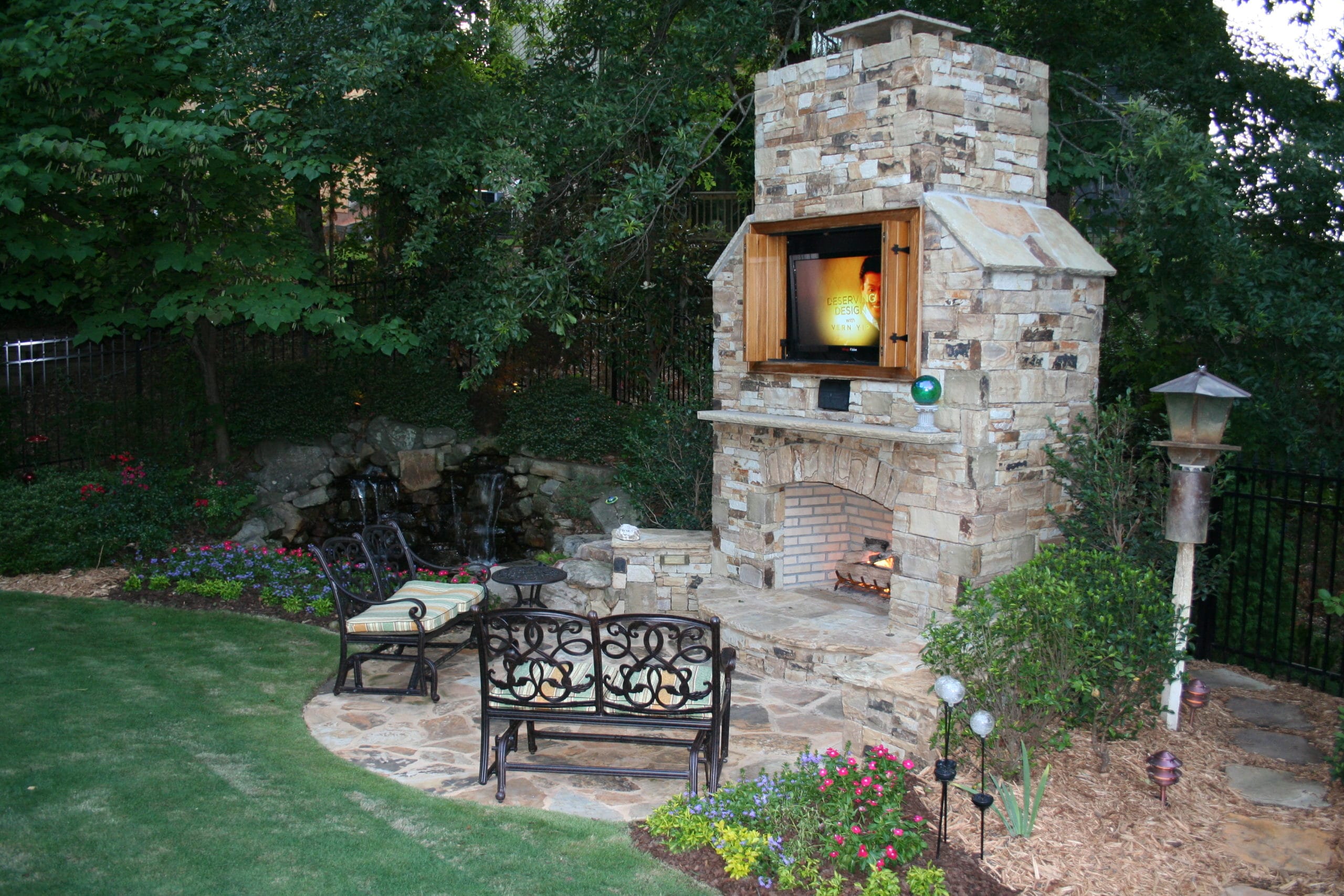 Outdoor stone fireplace with a mounted television, surrounded by wrought iron chairs and lush landscaping, including flowers, shrubs, and a small waterfall in the background.