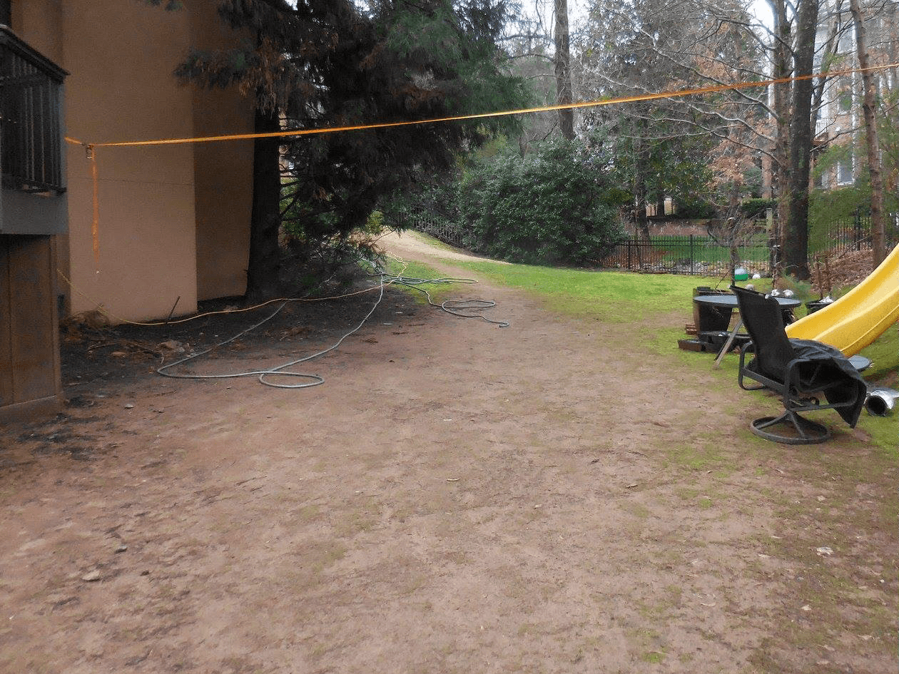 A backyard with patchy grass, a slide, chairs, hoses on the ground, and a yellow caution tape strung across the yard. Trees and shrubs line the background.