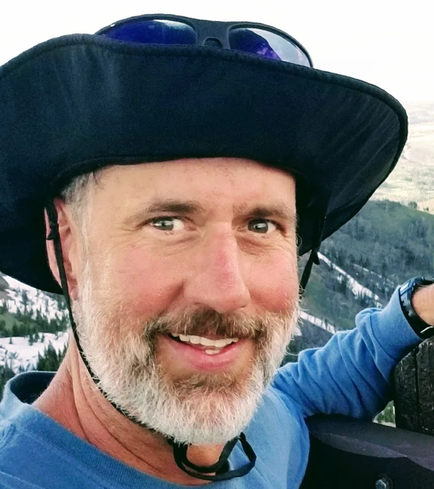 A man with a gray beard wearing a wide-brimmed hat and blue shirt smiles outdoors with mountains and trees in the background.