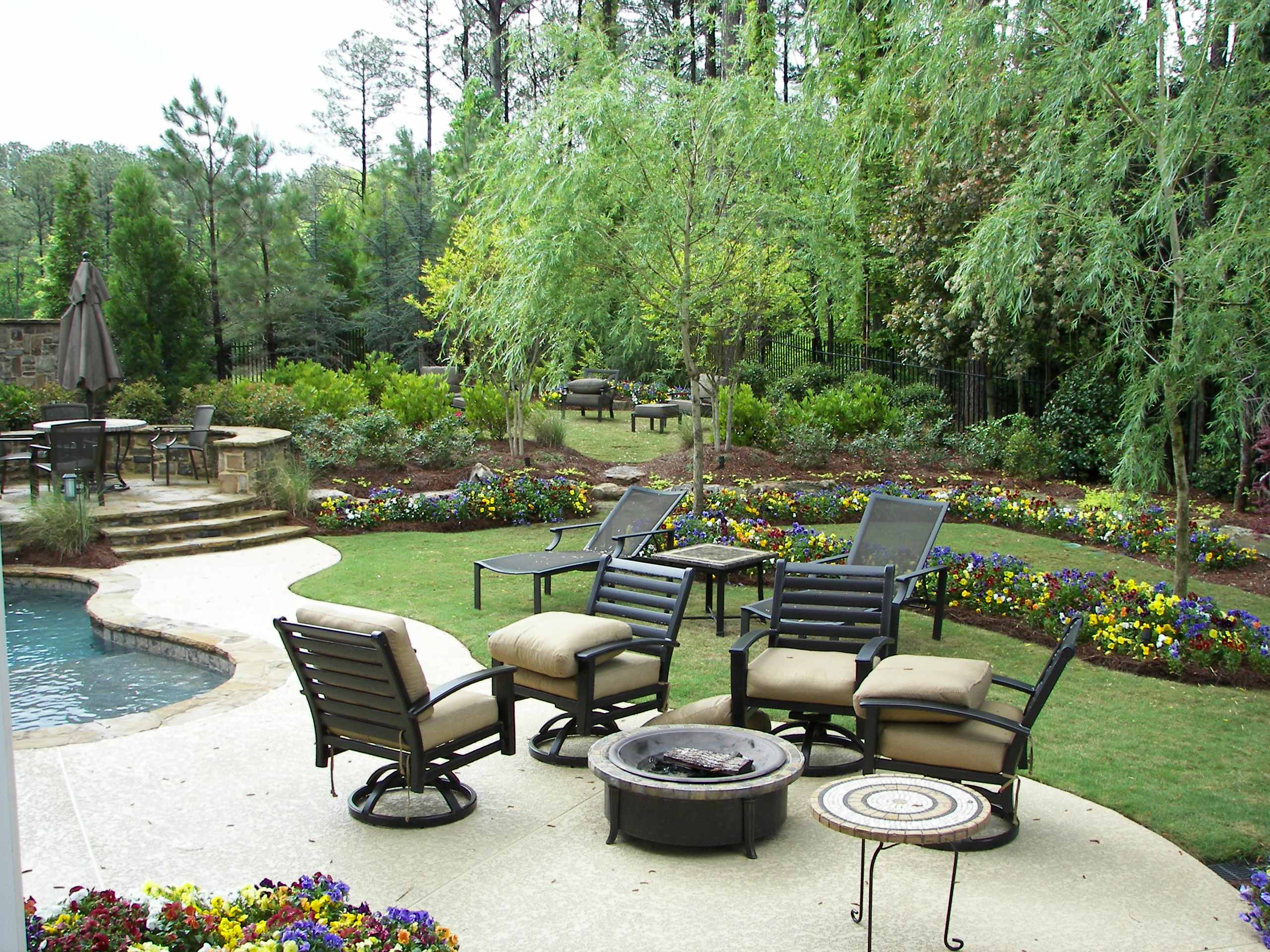 A landscaped backyard with patio furniture around a fire pit, colorful flower beds, a swimming pool, and trees in the background.