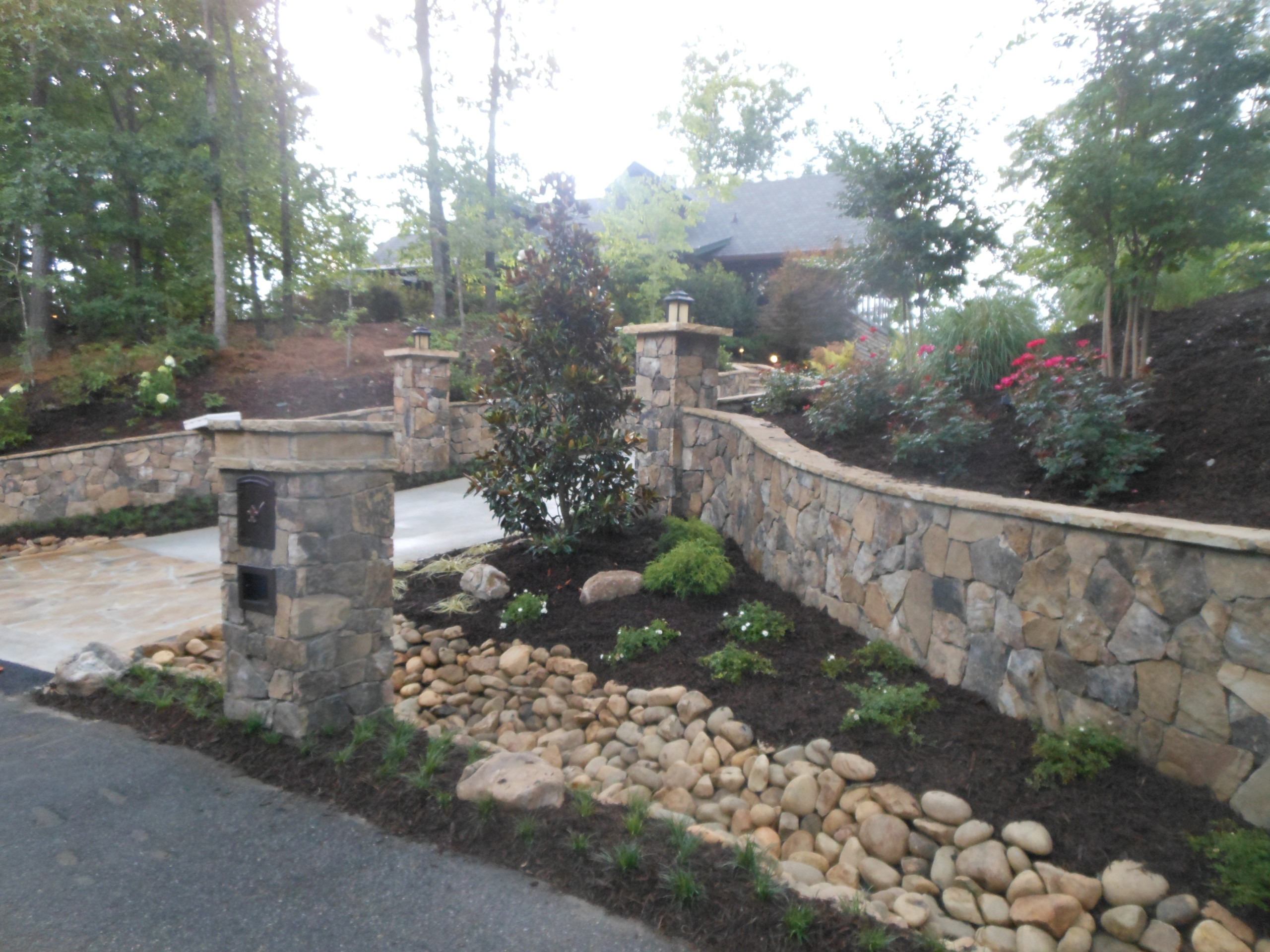Stone driveway entrance with mailbox pillar, landscaped garden beds, decorative rocks, and trees, leading up to a house partially visible in the background.