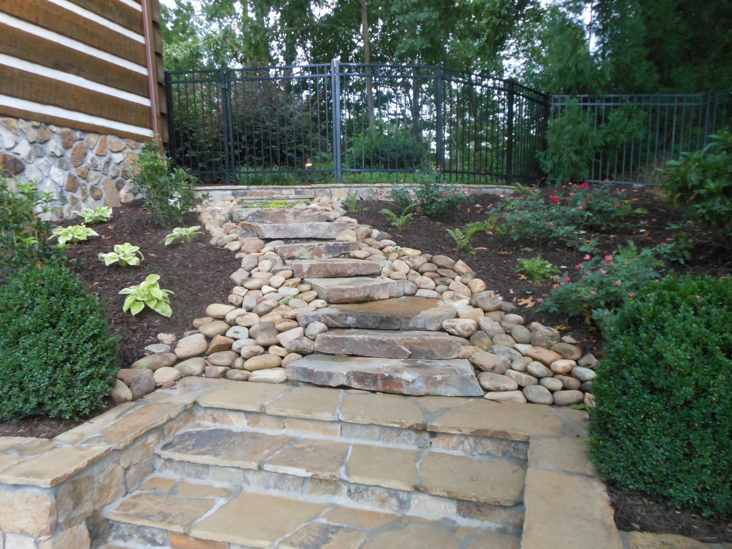 Stone steps lead up through landscaped garden beds with rocks and shrubs, ending at a metal fence and gate with trees in the background.