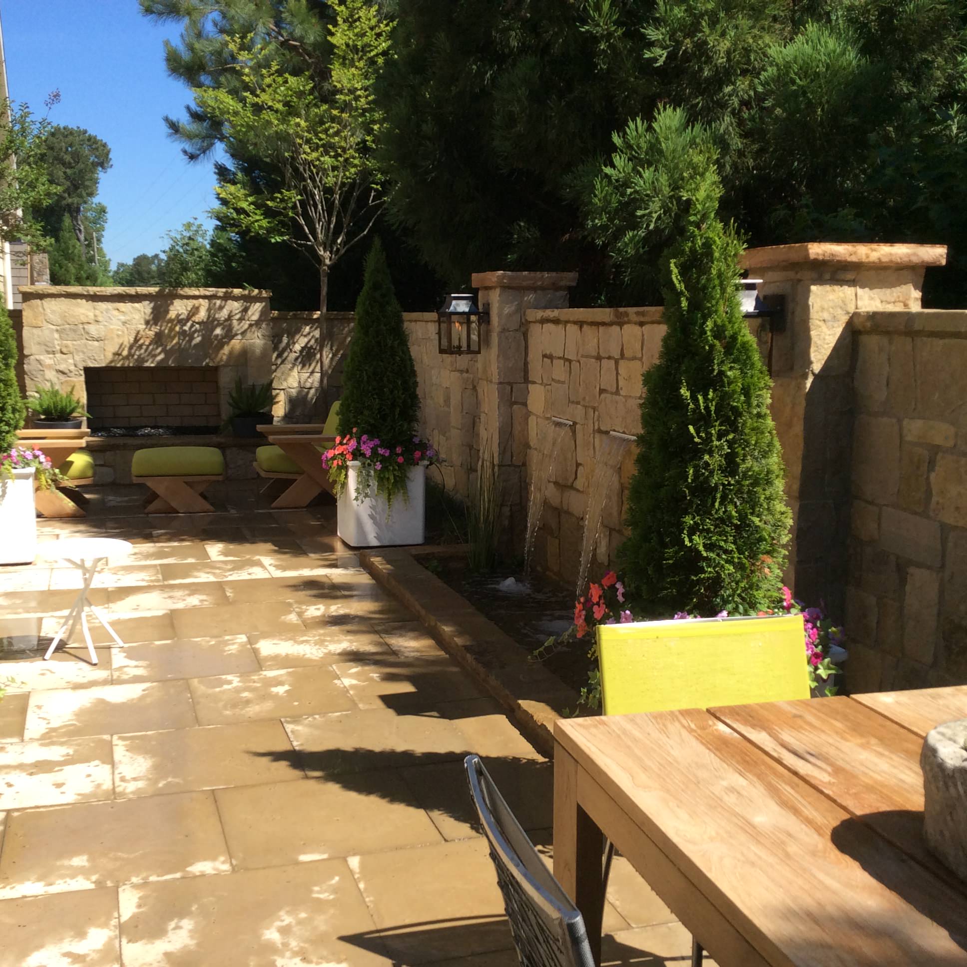 Outdoor patio with stone walls, potted plants, wooden table, yellow chairs, benches, and a brick fireplace under clear blue sky.