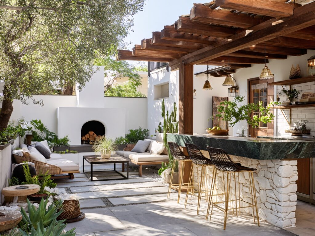 Cozy outdoor space featuring a pergola, bar seating, lounge area, fireplace, lush greenery, and modern decor in a Mediterranean-inspired setting.