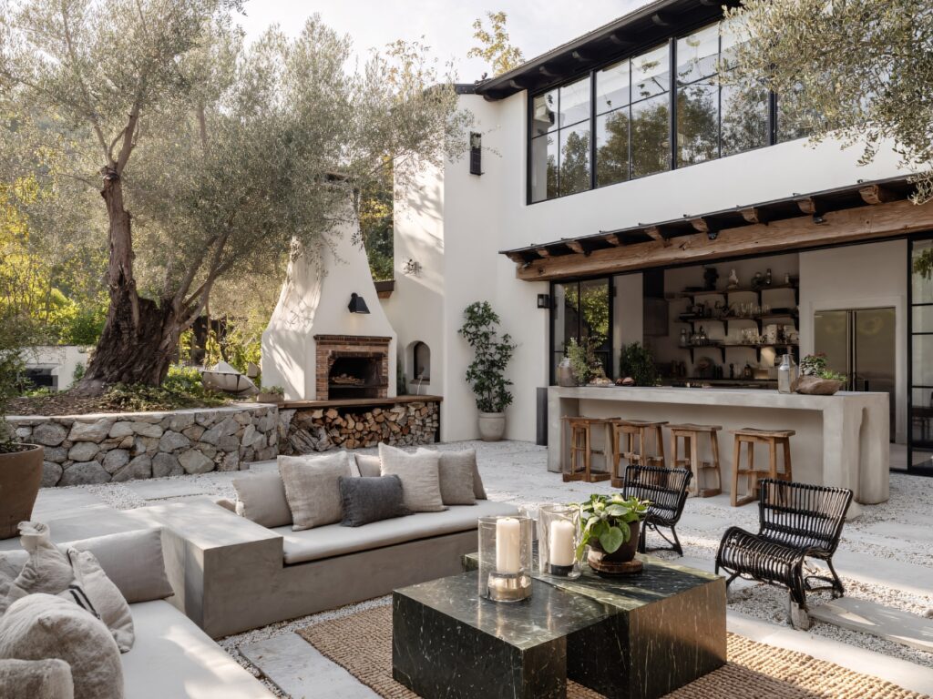 Modern outdoor patio creates a cozy outdoor space with a built-in sectional sofa, black chairs, marble coffee table, bar seating, open shelves, wood-burning fireplace, and stone landscaping beside the house.