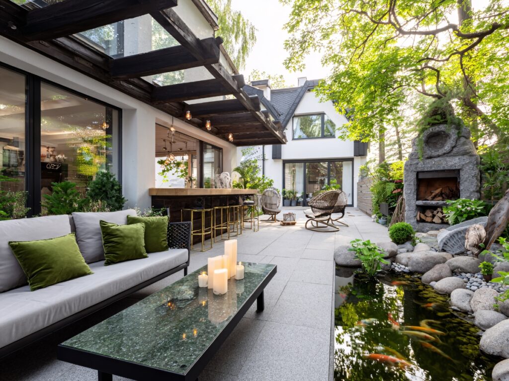 Modern patio featuring a cozy outdoor space with a sofa, green cushions, glass table with candles, bar seating, stone fireplace, and koi pond, all beside a contemporary house surrounded by lush trees and plants.