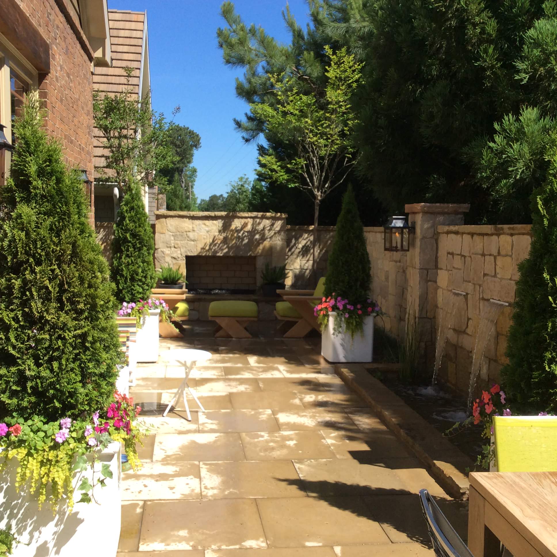 Sunny patio with stone paving, potted plants, benches with green cushions, and a stone wall lined with trees and shrubs.