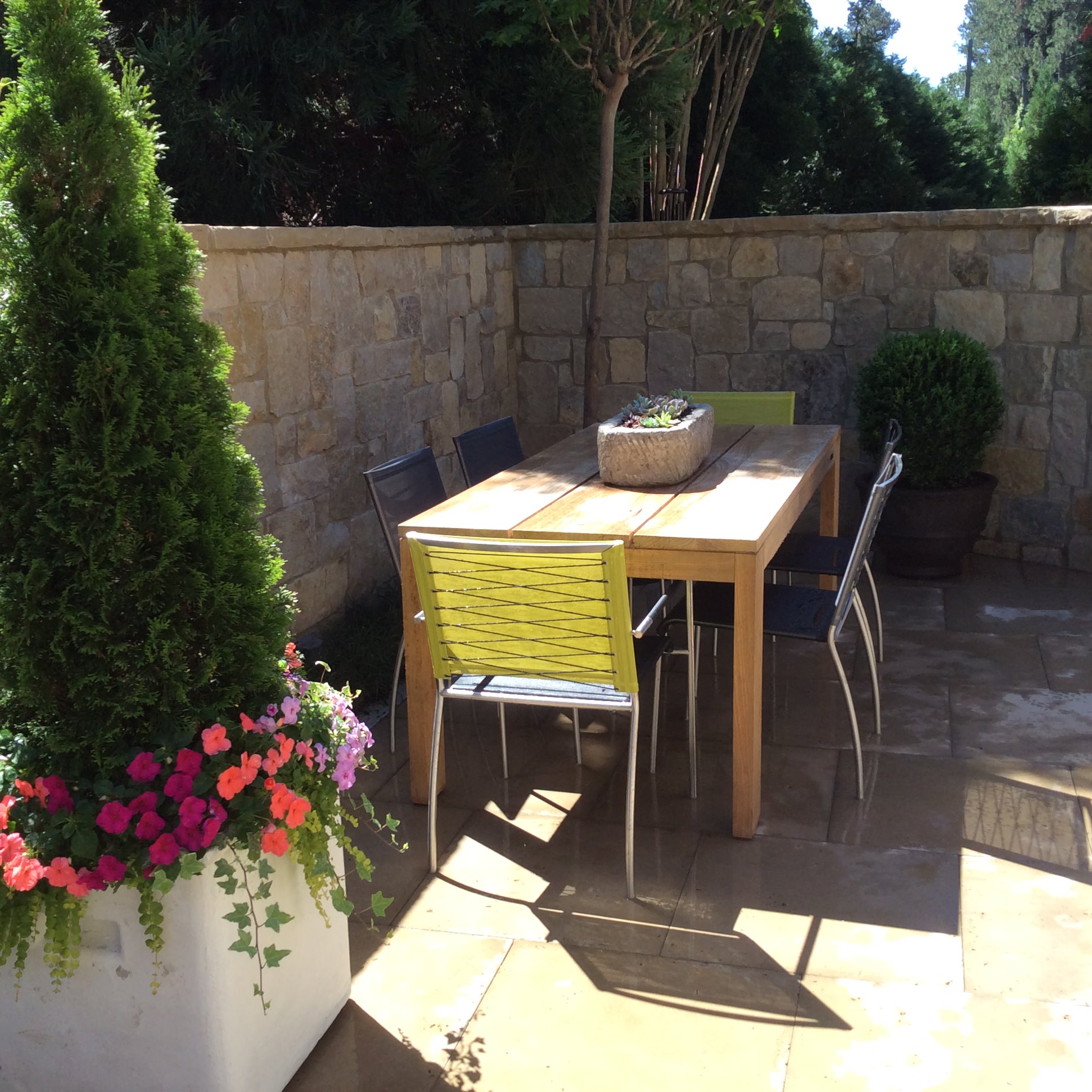 Outdoor patio with a wooden table, six chairs, potted plants, and a stone wall background on a sunny day.
