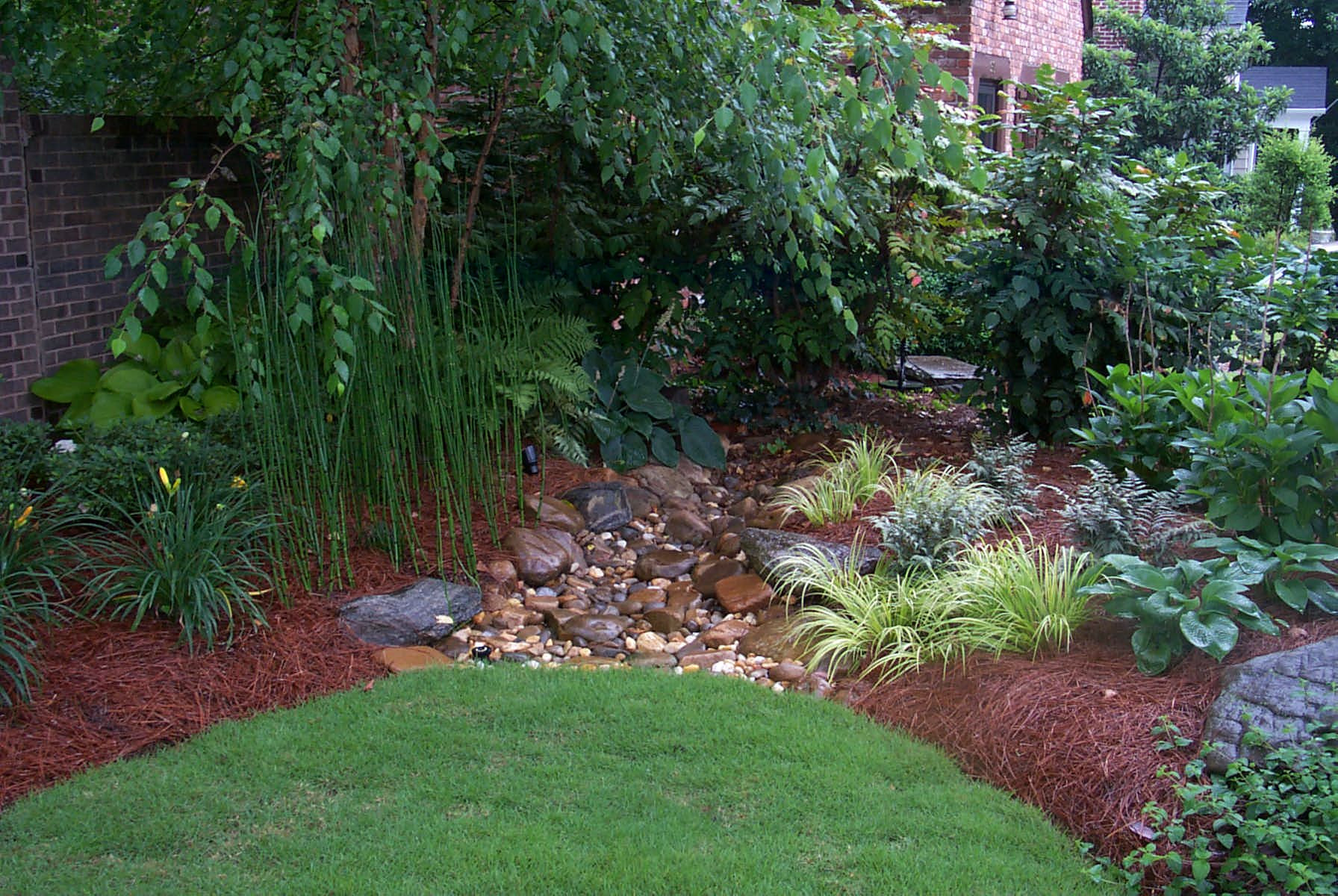 Landscaped garden with a small dry creek bed of rocks, surrounded by green plants, mulch, and grass near a brick house.