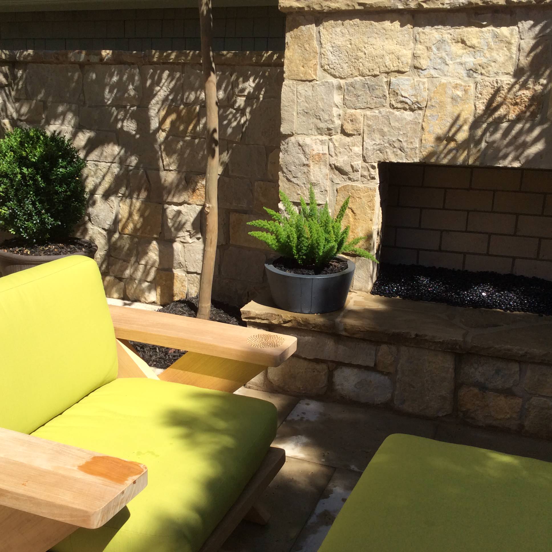 Outdoor patio with light green cushioned wooden chairs, a stone fireplace, a potted fern, and a manicured bush against a stone wall in sunlight.