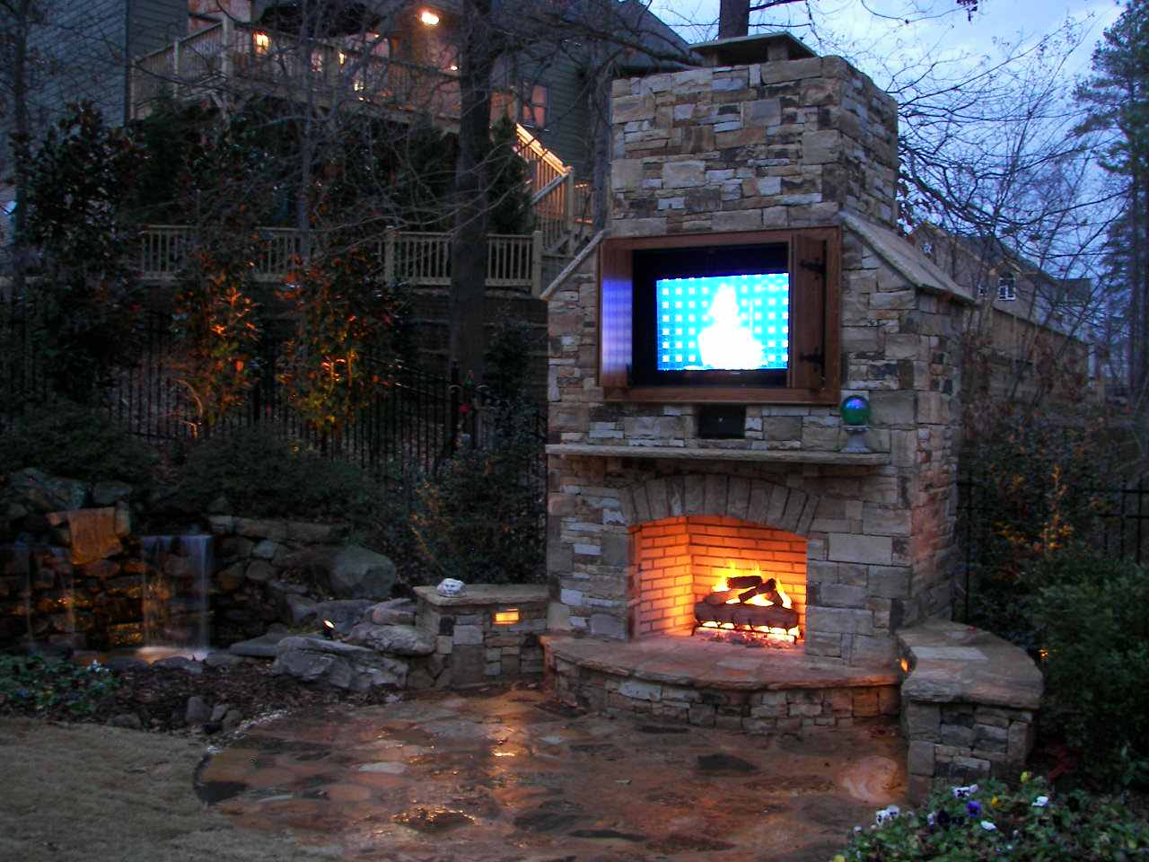 Stone outdoor fireplace with a lit fire beneath a mounted television, surrounded by a wet stone patio, garden, and waterfall feature at dusk.