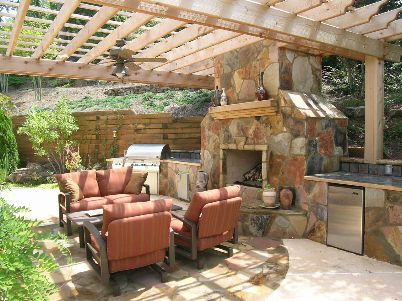 Atlanta Hardscapes Design: Outdoor patio with a stone fireplace, grill, mini fridge, and seating area under a wooden pergola.