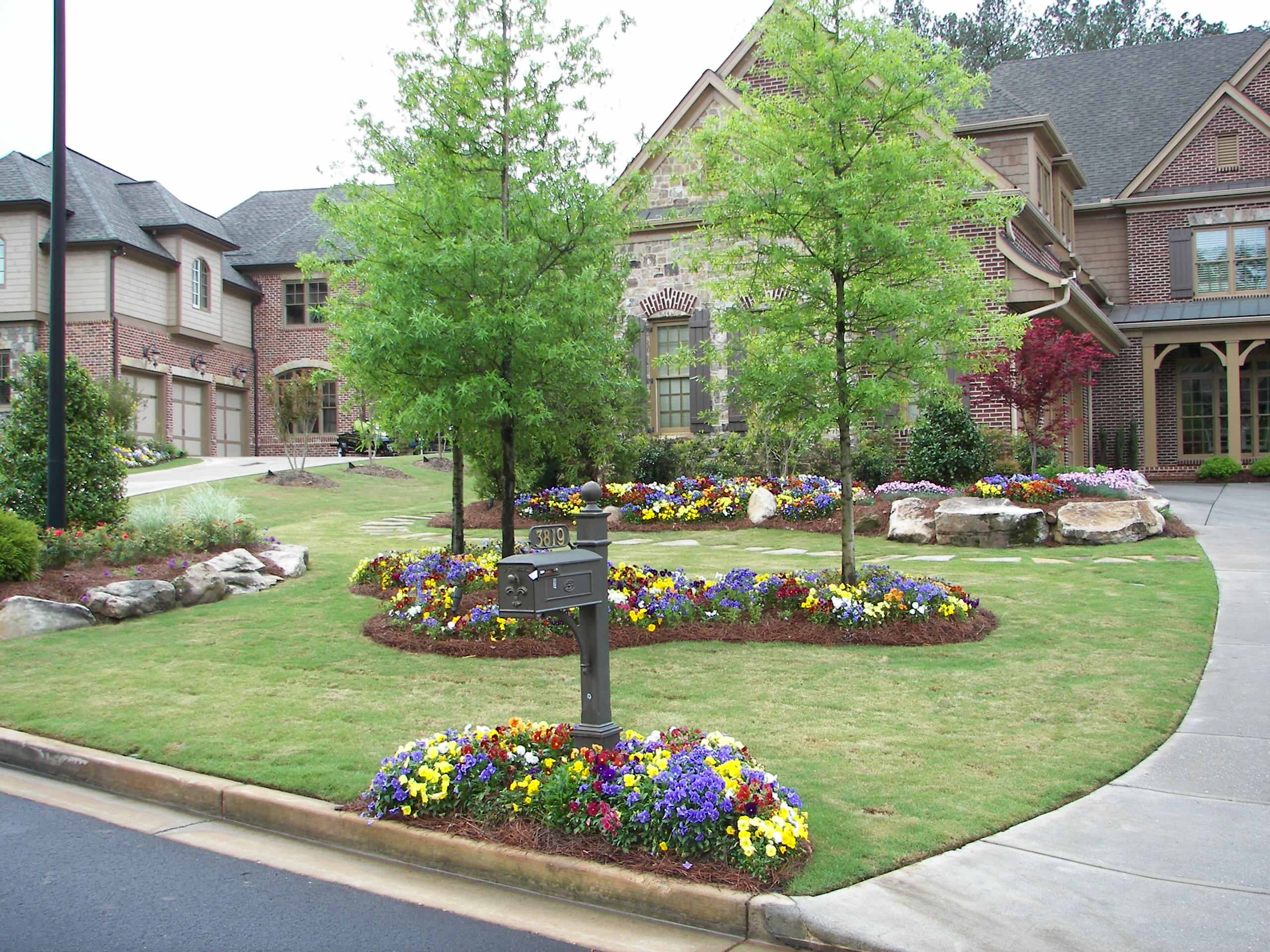 A neatly landscaped suburban front yard with colorful flower beds, trimmed grass, two trees, a sidewalk, and a mailbox, in front of large brick houses.