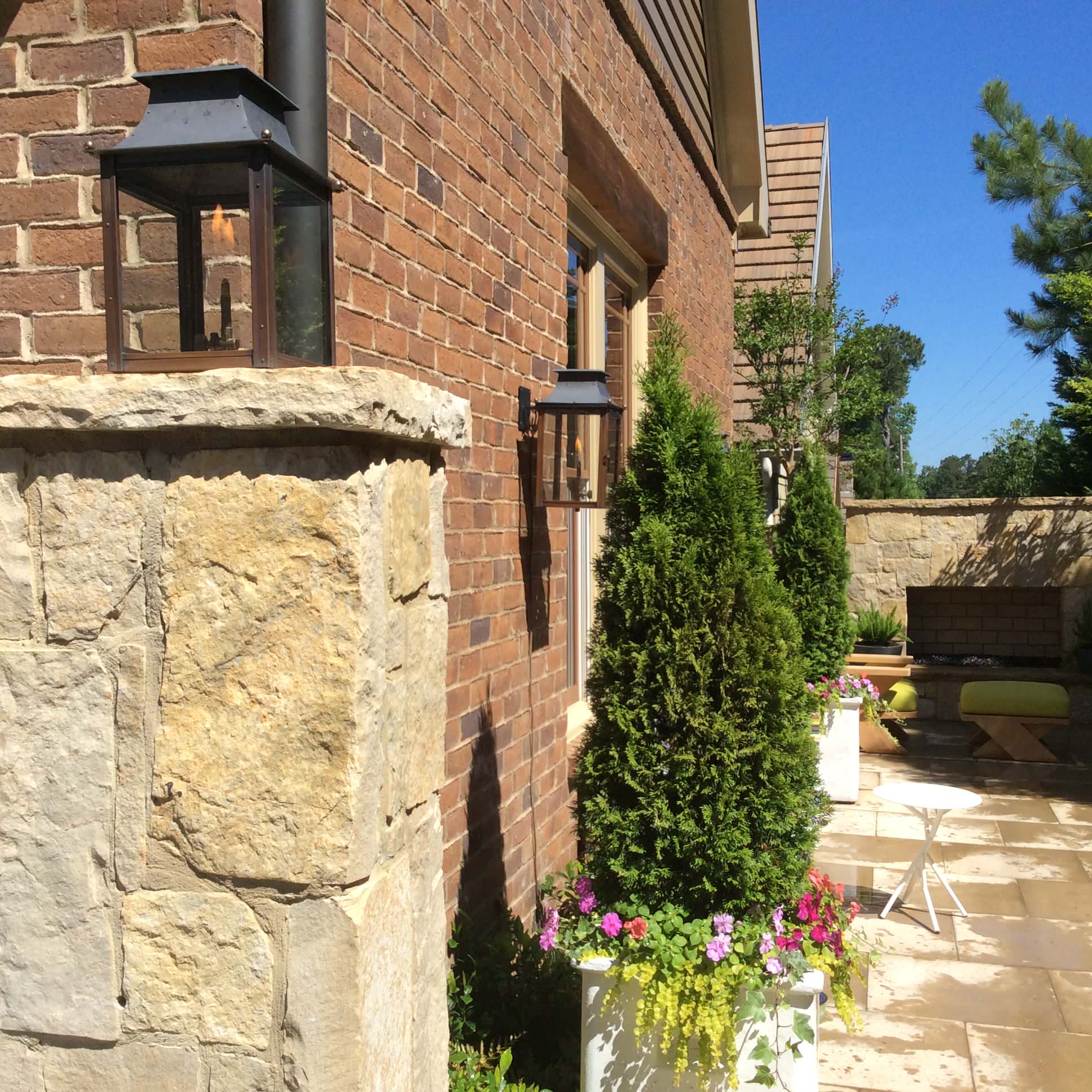 Brick house exterior with stone pillar, wall-mounted lanterns, potted evergreen trees, and flower boxes on a sunny patio.
